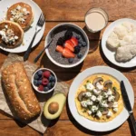 Low Carb Frühstück mit Protein-Pfannkuchen, Chia-Pudding, Omelett mit Pilzen und Feta, Nussbrot, Avocado und frischen Beeren auf einem rustikalen Holztisch.